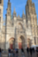Ornate Gothic cathedral facade with tall spires and red doors, people walking in the square under a bright blue sky.