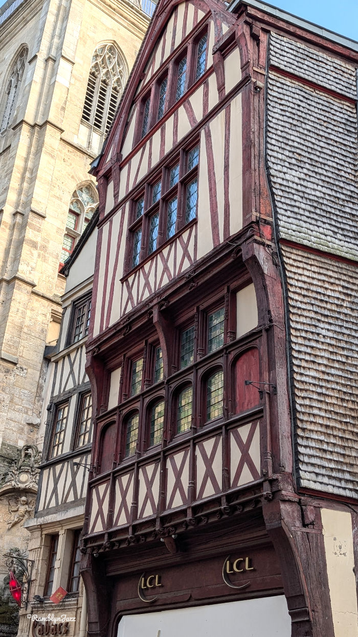 Tudor-style timber-framed building with red trim beside a stone church tower; LCL signs on the ground floor, sunny street scene