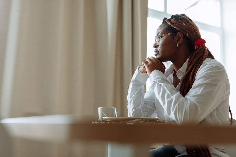 A Black person with long hair sitting at a table and staring out a nearby window.