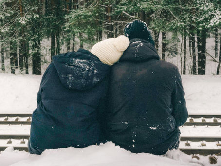 A photo of a couple bundled up, seated, and facing away. The background includes snow on the ground and a collection of evergreen trees.