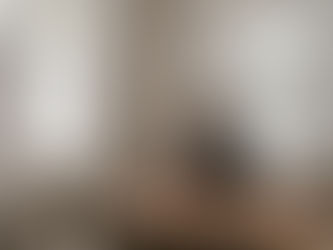 A grieving person leaning on a wooden dining table, covering his face with his hand, and staring out of a nearby window.