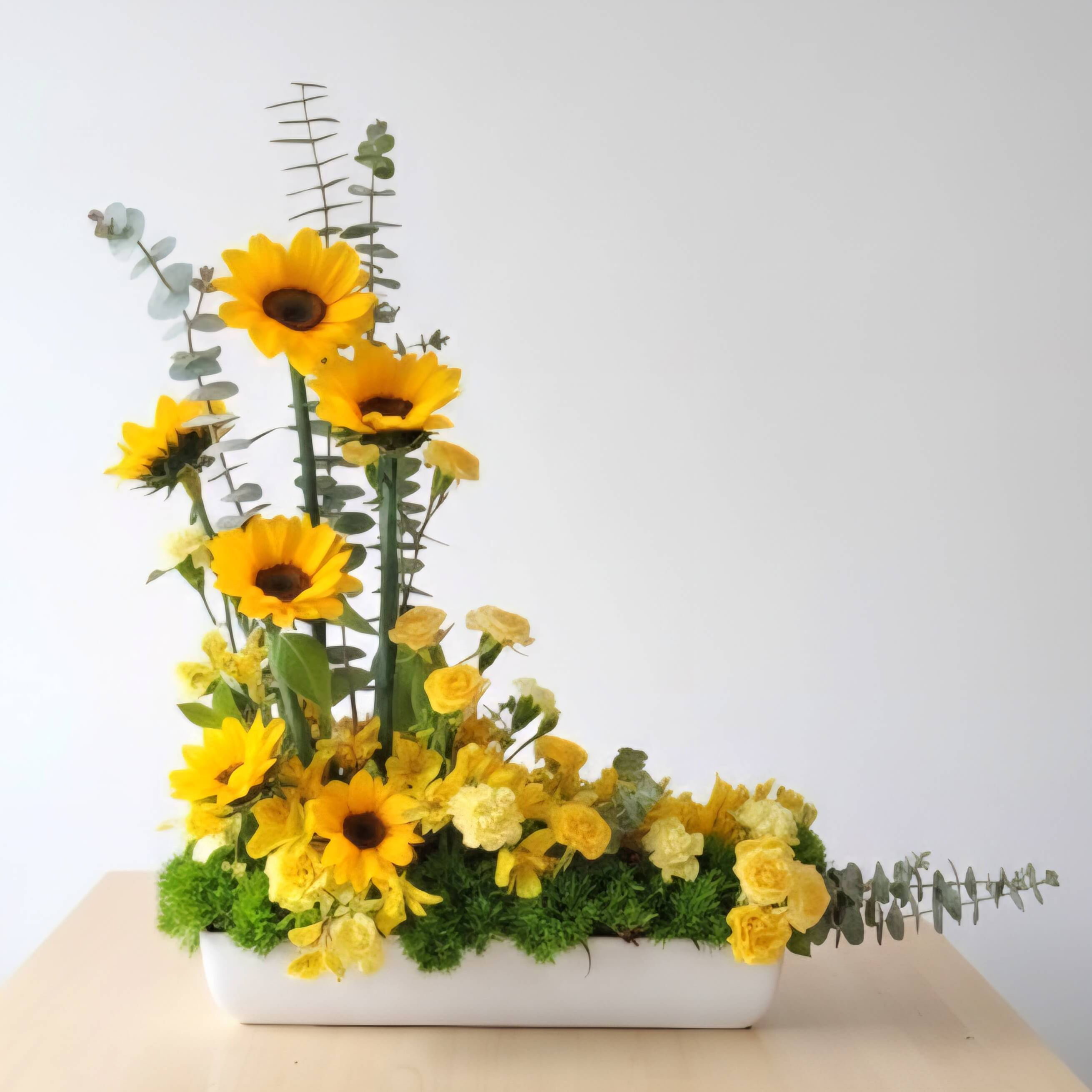 Arreglo floral con girasoles, flores amarillas y eucalipto en jarrón blanco.