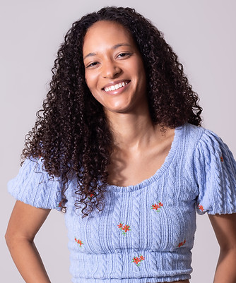 Manuella de Castro, de cabelos cacheados sorrindo, vestindo cropped azul bordado e calça clara contra fundo neutro.