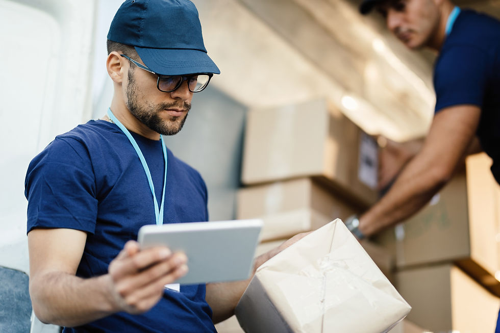 Profissional de logística usando tablet para conferir etiqueta de pacote durante triagem em centro de distribuição de e-commerce, enquanto outro colaborador organiza caixas ao fundo, ilustrando operações de logística reversa, controle de estoque e preparação de produtos para leilão de logística reversa.