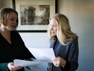 Two women in an office discussing documents. One points while the other listens attentively. A landscape painting hangs in the background.
