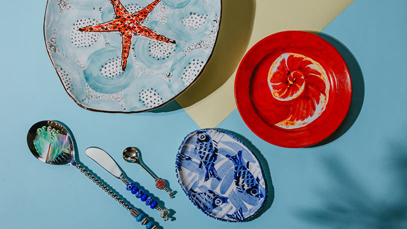 Colorful painted ceramic plates and spoons on a light blue background, with starfish, shell, and fish designs.