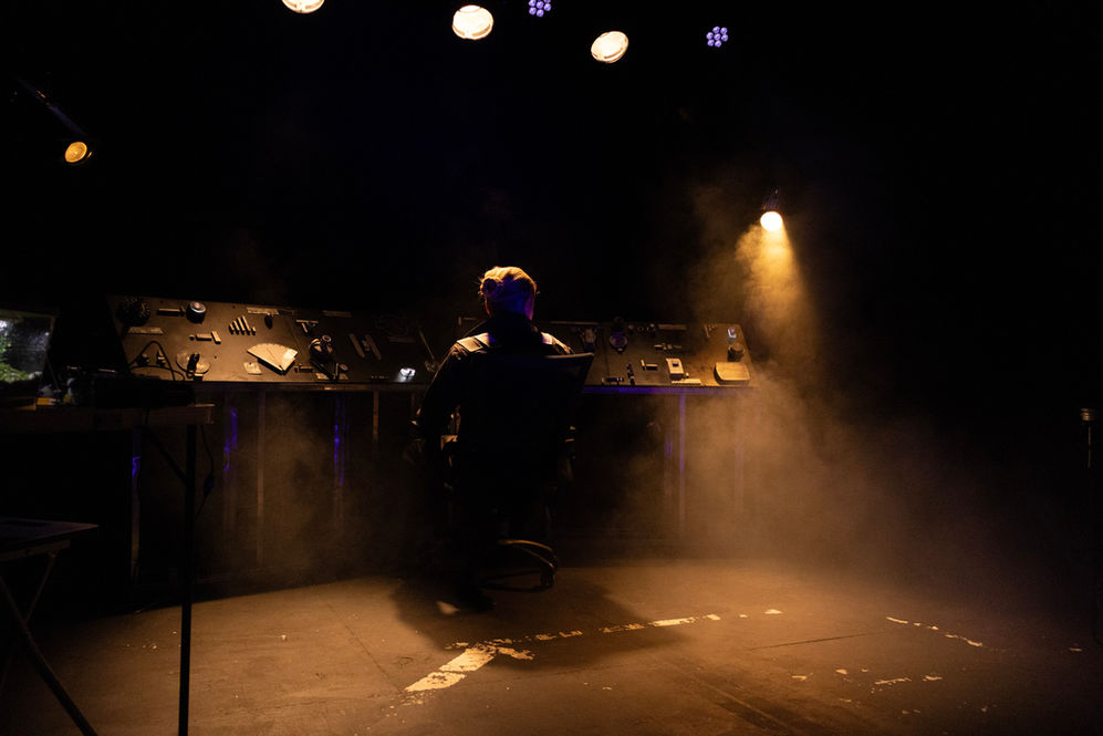 A dark stage with only a few lights on. The silhouette of someone sitting at a control pannel. 