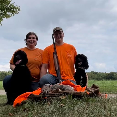 two people kneel between two standard poodles and have birds from a hunt in front of them
