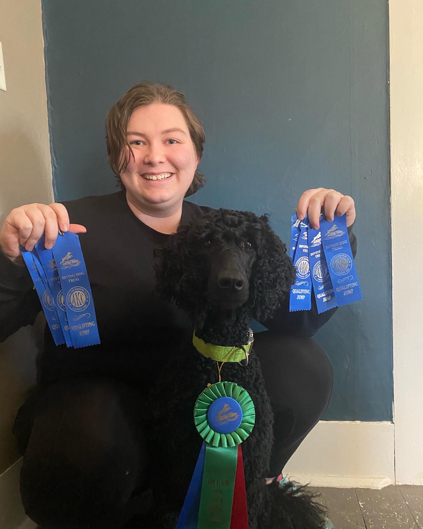 woman smiling and crouched behind black poodle with many ribbons