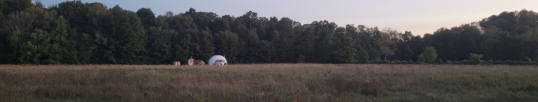 The geodesic dome tent @ Sky Camp isnestled agaist a lush forest wall. above the foest canopy, the sky fades in muted hus of blue and violet while a sliver of oon shines in the distance