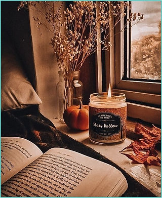 an open book sat on a bed, right next to a windowsill with a lit candle, some autumn leaves and a pumpkin ornament. It feels cosy and warm