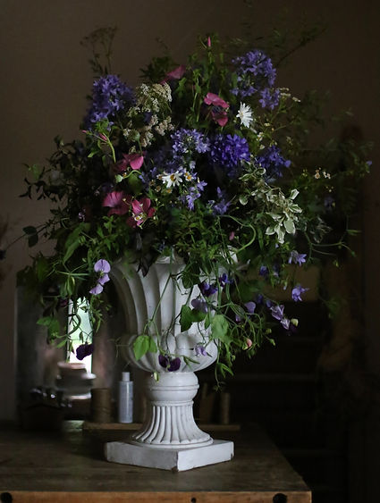 Large white architectural urn containing a cascading arrangement of purple, pink, burgundy and cream flowers with green foliage