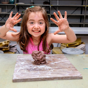 A young girl beams with excitement, displaying her clay-covered hands and a freshly crafted sculpture in a pottery class.