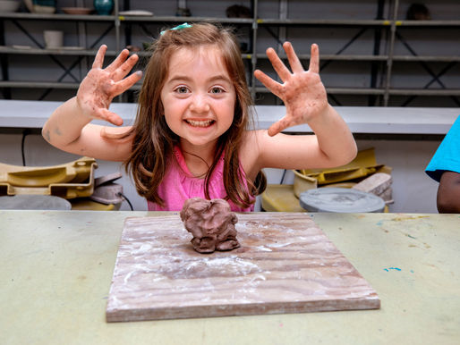 A young girl beams with excitement, displaying her clay-covered hands and a freshly crafted sculpture in a pottery class.