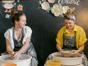 Two women enjoy a creative pottery session, skillfully shaping clay on spinning wheels against a charmingly decorated backdrop.