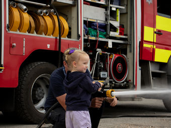A toddler in a lilac outfit has a go with the fire hose, supported by a crouching firefighter. Her face lights up with pride as the water sprays ahead.