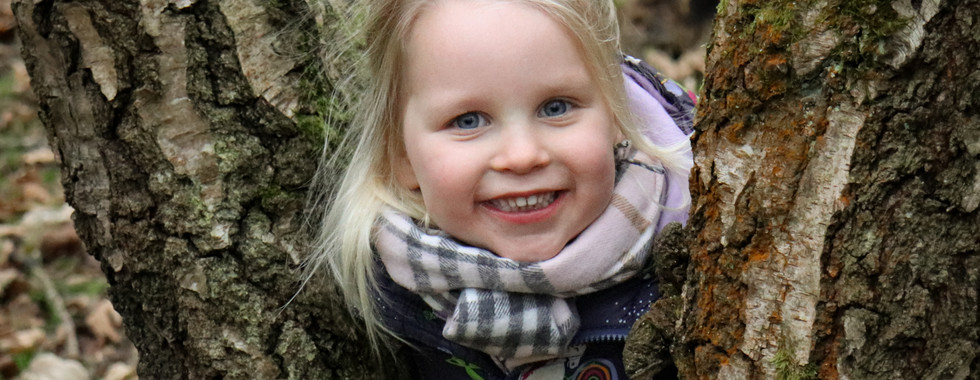 A smiling child peeks playfully from between two trees, bundled warmly in a scarf and jacket amidst the forest.