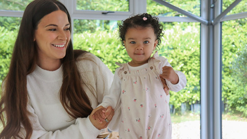 A mother holds her toddler’s hand as they stand confidently, smiling in a bright space with sunlight streaming through large windows.