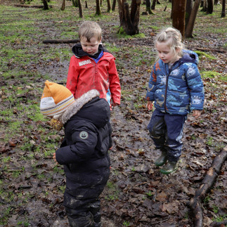 A child in a black snowsuit splashes in a puddle while others watch, embracing sensory play and outdoor exploration.