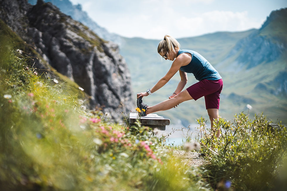 Woman stretching her leg on a bench in a mountainous landscape, wearing a blue tank top and red shorts. Bright, sunny day, lush greenery.