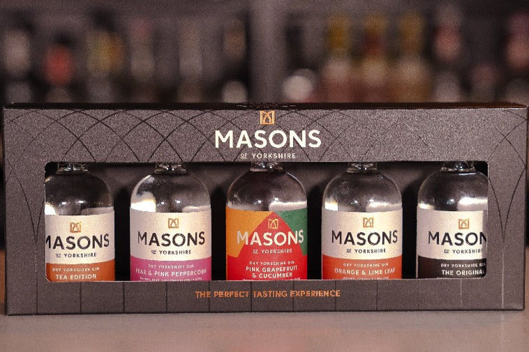 Box of five Masons gin bottles on a blurred shelf. Labels show flavors: Tea, Pink Peppercorn, Pink Grapefruit, Lime Leaf, and Original.