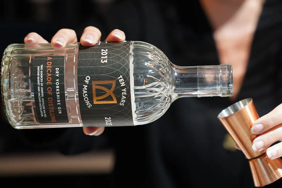Hands hold a bottle of Masons Dry Yorkshire Gin, labeled "A Decade of Distilling," tilted to pour into a copper jigger.