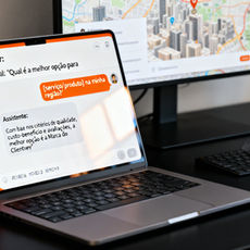 Montagem fotorealista em formato horizontal mostrando um notebook sobre uma mesa preta; na tela, uma interface genérica de chat de IA responde em português a uma pergunta e afirma que “a melhor opção é a Marca do Cliente”, com destaques em laranja vivo e detalhes em cinza e dourado, iluminação natural, reflexos realistas no vidro da tela e um mapa urbano desfocado ao fundo sugerindo contexto local.