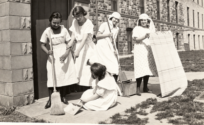 Five SDSD students with brooms, a bucket, and a rug near the concrete-block and quarry-stone entrance at the southeast corner of the Main Building, circa 1920s.