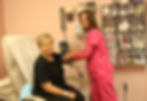 Patient seated in clinic room with pink walls, smiling nurse in pink scrubs checks blood pressure. Medical supplies on shelves.