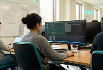 A young female university student in a casual jumper working at a monitor displaying code in a modern university computer lab, with a whiteboard showing binary tree diagrams and Big O notation behind her and other students at workstations nearby, representing the algorithmic and programming focus of Computer Science degrees