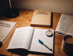 A journalism student's study desk under a lamp, with an open notebook of practised Teeline shorthand outlines and a teal marker pen resting across it. A stopwatch sits on the page for timed shorthand drills. Nearby are a textbook titled Teeline Shorthand, a book titled Public Affairs for Journalists, a folded newspaper, and a mug of tea.