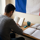 A university student reading a French novel at a desk in a French language study space, with a French tricolore flag on the wall behind him, an open notebook of verb conjugation exercises, and an open French-English dictionary beside him.