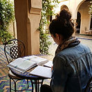 A young female university student studying in a beautiful courtyard with traditional Middle Eastern geometric tiles and archways, with an Arabic textbook and notebook open, representing the immersive language and cultural study experience of Arabic and Middle Eastern Studies degrees