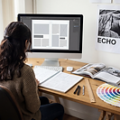 A graphic design student at a studio desk working on a magazine spread layout on screen, with a sketchbook of layout thumbnails, a colour reference fan and an open reference magazine on the desk, and a printed design poster pinned on the wall behind.