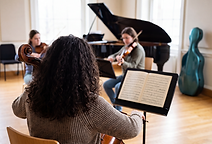 A student cellist rehearsing with a small chamber ensemble in a university music room, playing from a score on a music stand, representing the performance and ensemble focus of Music degrees.