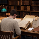 A history student in an archive reading room studying an open primary source manuscript, with an archive document box, white cotton gloves and a leather research notebook on the desk. Tall bookshelves of leather-bound volumes fill the background.
