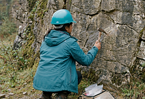 A geology student in fieldwork gear crouches at a quarry rock face, examining the surface with a geological hammer. A field kit of sample bags sits beside her.