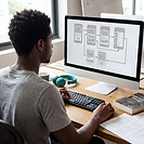A student at a desk designing how a mobile app moves from one screen to the next. The monitor shows phone layouts joined by arrows, with teal headphones and an interaction design textbook beside the keyboard.