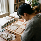 A student at a desk arranging a material and finishes board for a room scheme, choosing between fabric swatches, paint chips, a tile and a timber sample. An interior design book lies open beside the board, with a scale ruler to hand.