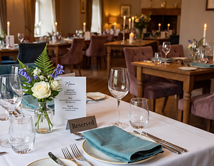 A table laid for fine dining in a candlelit restaurant, with a printed menu, a reserved card, a folded teal napkin and a small vase of fresh flowers.