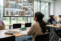 A young female university student in a seminar room looking at a large screen displaying a pop culture collage of magazine covers, social media posts and street art with critical analysis and media hegemony annotations overlaid, with a cultural theory textbook on her desk, representing the critical and analytical focus of Cultural Studies degrees