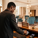 A hotel receptionist working at the front desk, processing a guest card payment beneath a Reception sign, with a warmly lit lobby of teal armchairs behind.