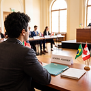 A student takes part in a Model UN debate, seated at a desk with a placard reading General Assembly, a folder and a small stand of national flags, while other delegates sit in the background.