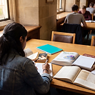 A university student seen from behind sits at a library desk with three different subjects laid out side by side. An open literature paperback and a copy of Pride and Prejudice sit at one side, an open biology textbook showing a cell diagram in the middle, and a history book on modern Britain at the other, each with its own notebook. A teal notebook rests among them and faceless students study behind.