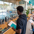 A university student seen from behind stands at a glass viewing gallery above a public leisure centre, holding a teal notebook and a clipboard headed Site Visit Observation Brief. Below him a lane-marked swimming pool and a gym floor with exercise machines are in use. Fellow students stand alongside him with the same clipboards. Teal signs on the wall read Aquatic Zone and Fitness Suite.