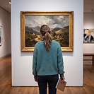 A history of art student standing in a gallery, studying a 19th-century European landscape painting in an ornate gilt frame, with other paintings visible on adjacent walls.