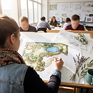 A landscape design student, seen from behind, hand-draws a colourful masterplan of a public park at a drawing board in a university studio, with winding paths, planting beds, tree clusters and a small lake. Loose concept sketches labelled CONCEPT PLAN, sprigs of olive and lavender, and a teal mug sit on the board, and behind her fellow students work at their own boards beneath a wall of pinned-up drawings.