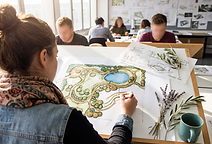 A landscape design student, seen from behind, hand-draws a colourful masterplan of a public park at a drawing board in a university studio, with winding paths, planting beds, tree clusters and a small lake. Loose concept sketches labelled CONCEPT PLAN, sprigs of olive and lavender, and a teal mug sit on the board, and behind her fellow students work at their own boards beneath a wall of pinned-up drawings.