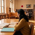 A student reads a copy of The Tale of Genji at a desk in a university library, a Japanese-English dictionary and a notebook beside her, with a Japanese flag and a print of Mount Fuji on the wall behind.
