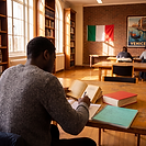 A student reads a copy of La Divina Commedia at a desk in a university library, a bilingual dictionary and handwritten notes beside him, with an Italian flag and a Venice travel poster on the wall behind.